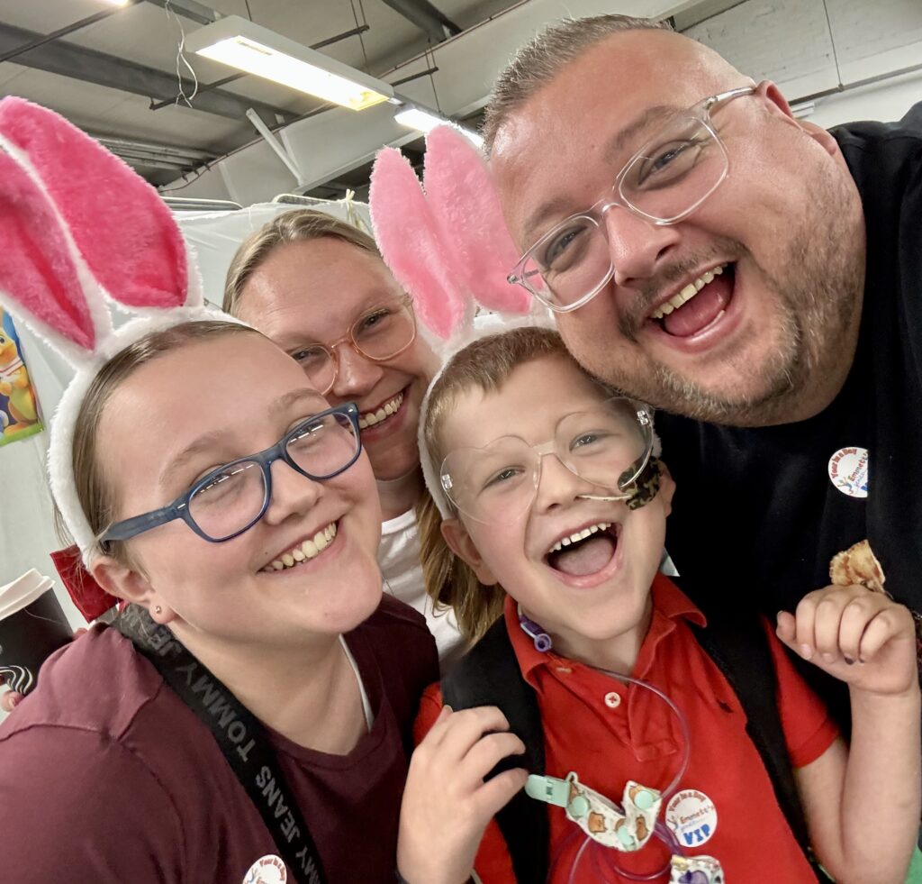A smiling family of four wearing bunny ear headbands pose for a selfie indoors. The child in front, part of Emmett's Genies, wears glasses and a red shirt. All are happy and close together, creating a joyful atmosphere.