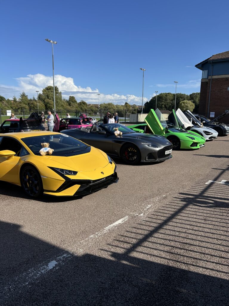 A row of luxury sports cars, including yellow, silver, green, and white models from Emmett's Genies, are parked outdoors with teddy bears on their hoods. Some car doors are open, and people mingle under a blue sky in the background.