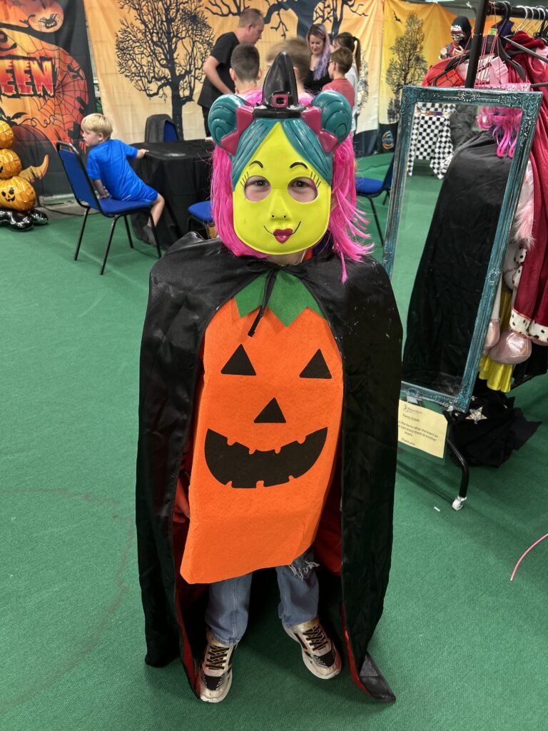 A child dressed in a colorful mask, witch hat, black cape, and pumpkin costume stands indoors on green carpet at Emmett's Genies Halloween event, with decorated pumpkins and other children visible in the background.