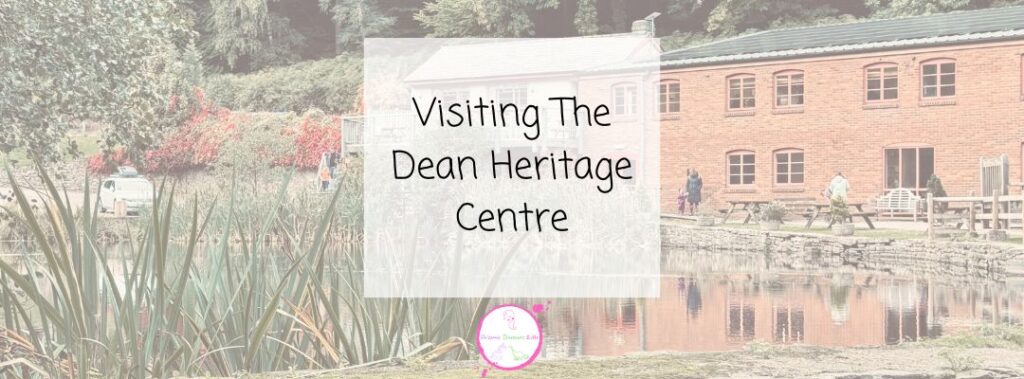 A pond with reeds in the foreground reflects the Dean Heritage Centre's brick building with large windows. People are walking and sitting outside. Text overlay reads: Visiting The Dean Heritage Centre.