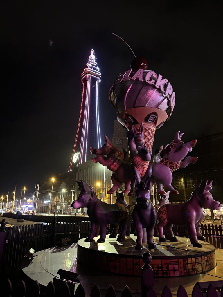 A night scene in Blackpool featuring the illuminated Blackpool Tower in the background and a whimsical sculpture of donkeys and an ice cream cone with the word BLACKPOOL on it in the foreground.
