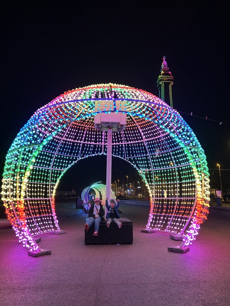 Two children sit on a bench under a large, colorful, illuminated tunnel structure at night, with vibrant rainbow lights and a tall, lit-up tower in the background.