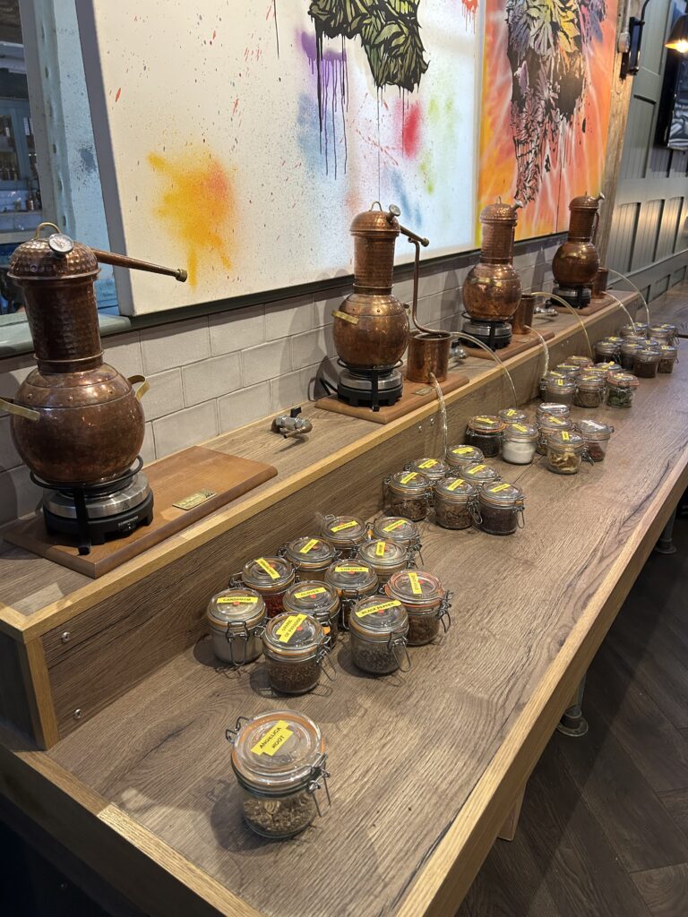 A wooden counter displays copper distillation apparatus and several glass jars filled with assorted spices and botanicals, each labelled, beneath colourful artwork on the wall.