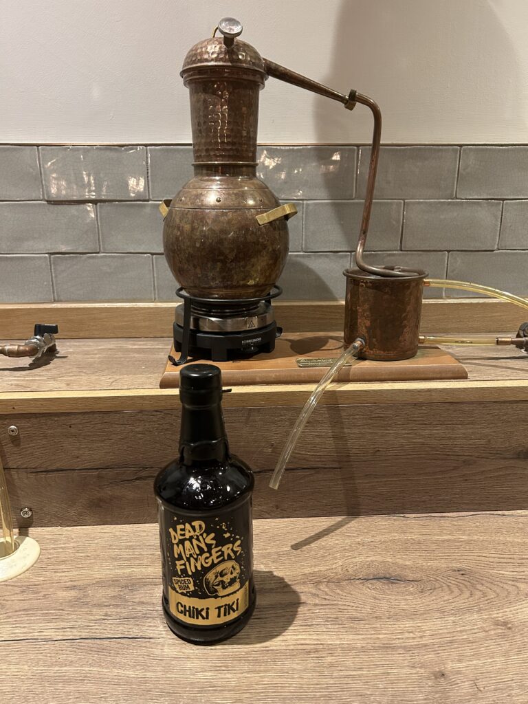 A copper still sits on a wooden counter with a bottle of Dead Man’s Fingers Chiki Tiki rum placed in front of it. The background features grey tiled walls and wood panelling.