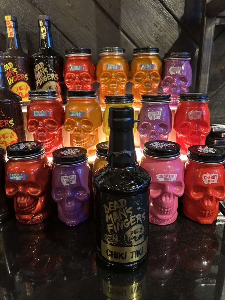 A display of colourful skull-shaped jars in red, purple, and orange, set behind a black bottle of Dead Man’s Fingers Chiki Tiki rum on a dark surface. The jars and bottle are illuminated from behind.