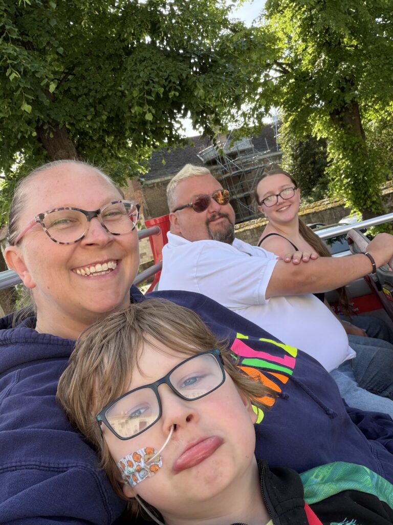 Four people, two adults and two children, smile whilst sitting on a tour bus. The child in front wears spectacles and a nasogastric tube on their face. Trees and a building are visible in the background.