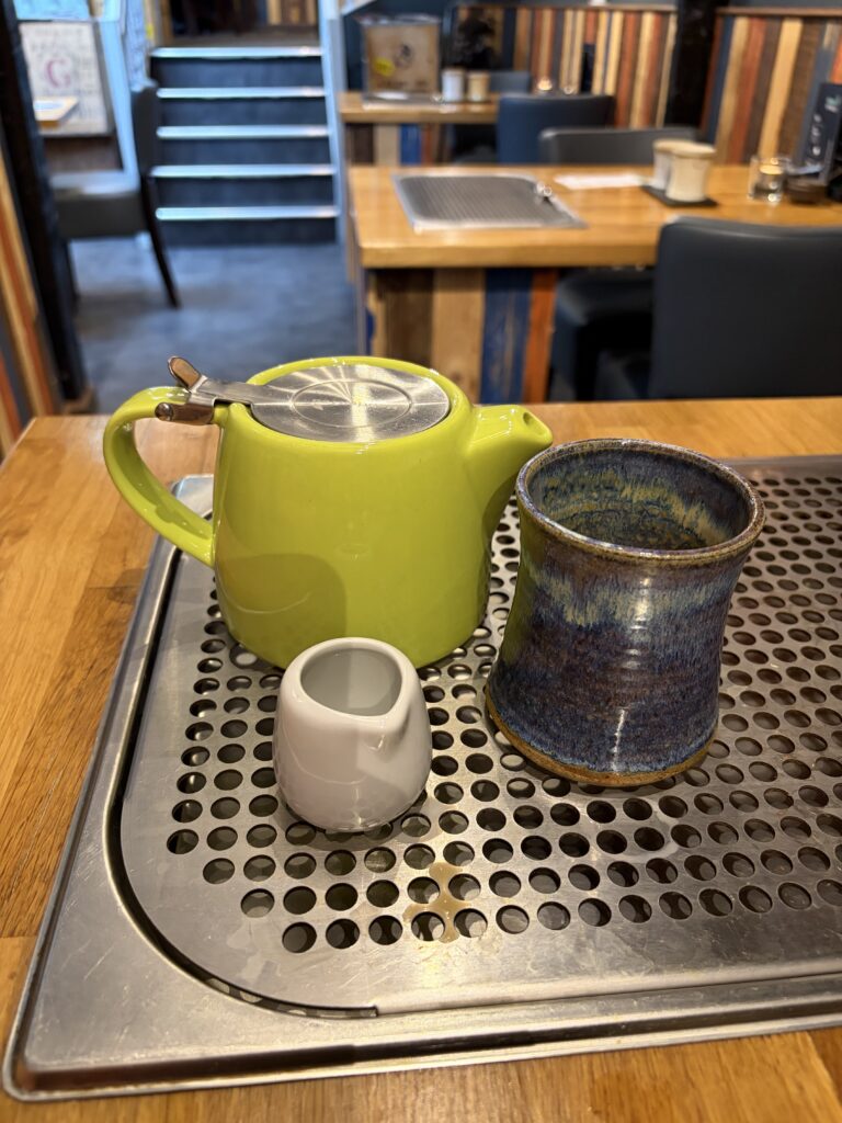 A green teapot, a blue ceramic cup, and a small white milk jug sit on a perforated metal tray on a wooden table in a cosy, colourful café.