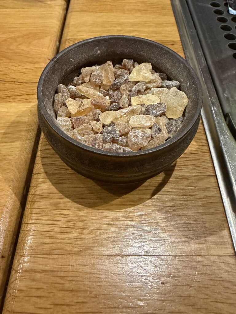 A brown ceramic bowl filled with irregularly shaped, amber and yellow sugar crystals sits on a wooden surface next to a metal grille.