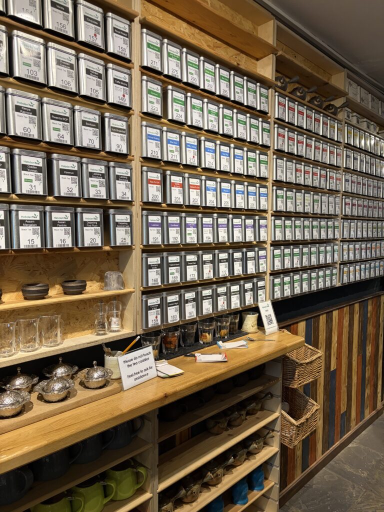 Metal canisters with labelled tags are organised on wooden shelves, displaying a variety of teas. Below, mugs, teapots, and sample glasses are neatly arranged on additional shelves and a wooden worktop.